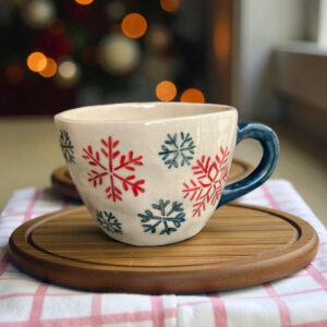 A handcrafted ceramic mug with red and blue snowflake patterns sits on a wooden coaster over a pink checkered cloth. Warm bokeh lights in the background create a cozy, festive winter atmosphere.