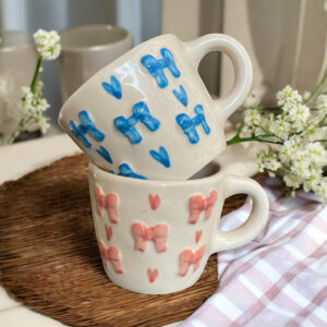 A set of 2 ceramic mugs , one with blue 3d bows and painted hearts and the other with pink 3d bows and hand painted hearts